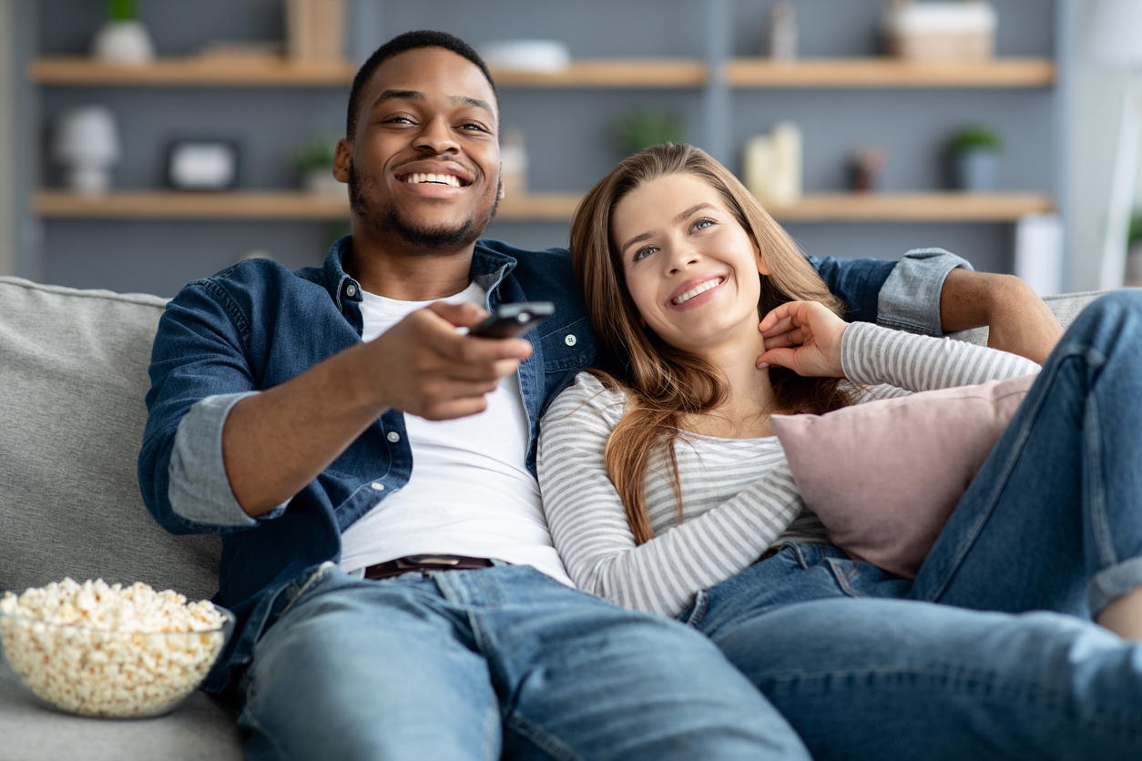 Couple sitting on couch with popcorn streaming Oppenheimer on TV