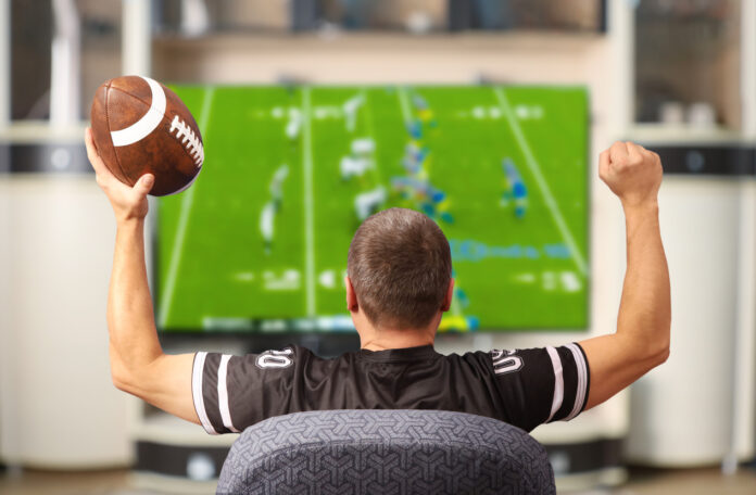 Fan of american football. Man with a ball watching TV. Man holding a football and wearing a football jersey cheers while watching a football game on TV