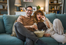 Man and woman sitting on the couch eating popcorn while streaming an action-packed movie at home.