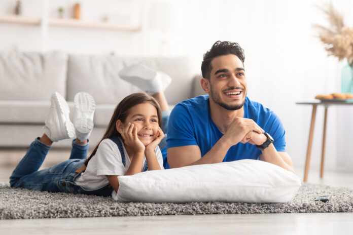 Happy young family watching television lying on floor Father and daughter lying on the living room floor while watching a movie together at home.